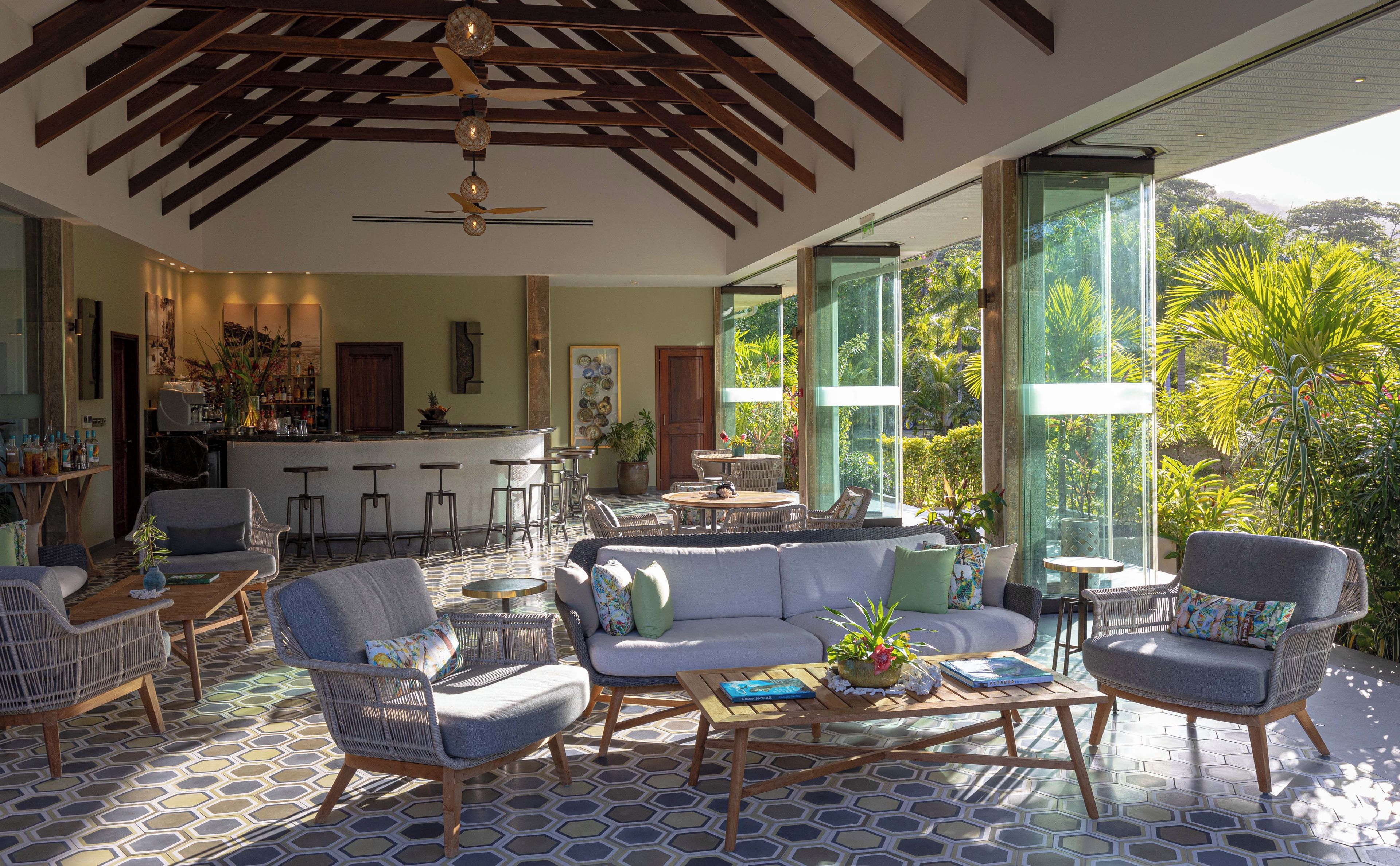 Amplia sala de estar con un sofá grande, dos sillones y una mesa de centro. Cuenta con una zona de cocina con barra y taburetes. La habitación tiene ventanas grandes que ofrecen vistas a un follaje exuberante.