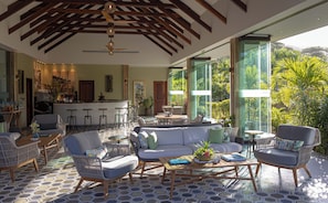 A spacious living area with a large sofa, two armchairs, and a coffee table. There is a kitchen area with a bar and stools. The room has large windows offering a view of lush greenery.