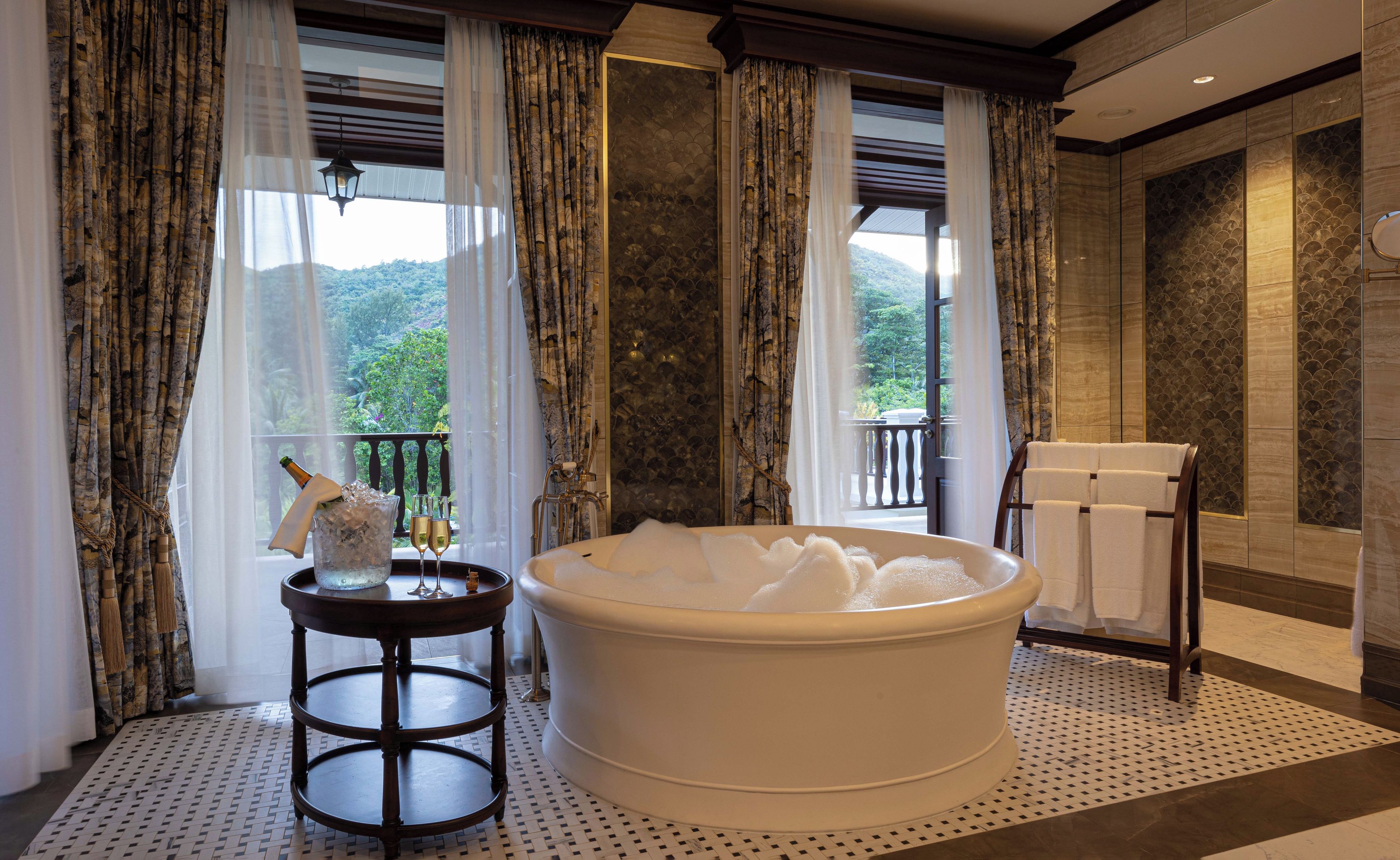 Ein Badezimmer mit einer großen Badewanne, einem kleinen runden Tisch mit einer Flasche und Gläsern sowie Blick ins Grüne.