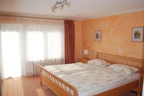 Uma cama de casal com colcha florida, uma mesinha de cabeceira com abajur e uma janela com cortinas.