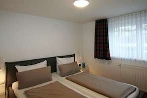 Quarto de hotel com duas camas, uma janela com cortinas, um radiador e uma luminária fixada na parede.