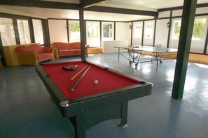 A pool table with a red felt surface, a billiards cue, and a white cue ball. There is a sofa with orange cushions and a table tennis table in the background. - Holiday apartment with 1 bedroom (Willingen-Usseln)