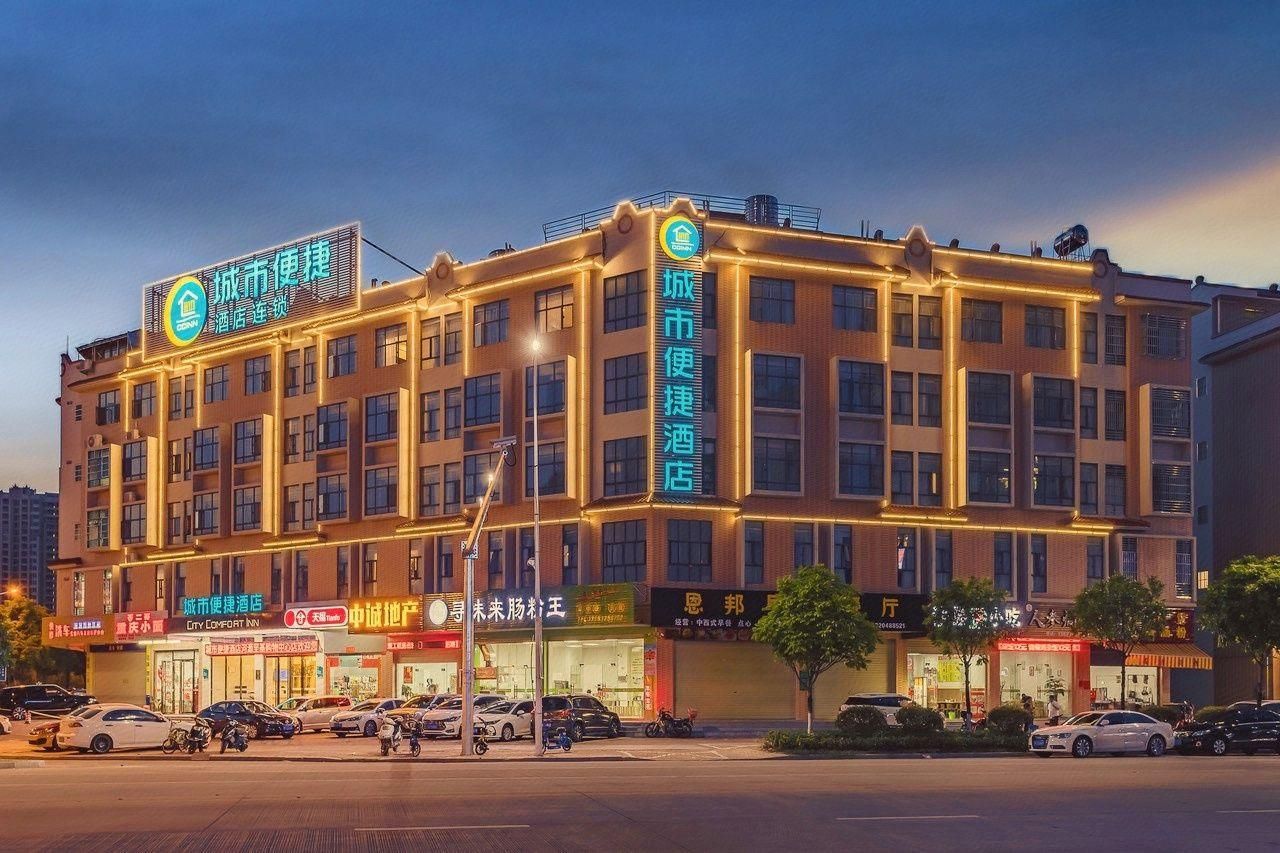 Un hotel de varios pisos con señalización iluminada, un estacionamiento con varios coches y diversas tiendas en la fachada.