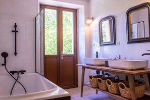 A bathroom with a bathtub, two sinks, a mirror, and a window with a view of greenery. - Holiday home in Treserve with Wi-Fi (Tresserve)