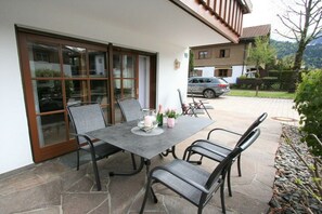 Um pátio externo com mesa e cadeiras, uma porta de vidro e vista para carros e edifícios ao fundo.