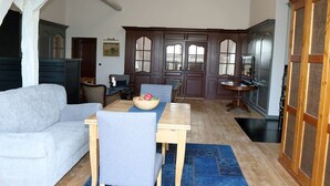Uma sala de jantar com uma mesa de madeira, cadeiras azuis e uma tigela de frutas. Há um sofá branco e um armário de madeira escura ao fundo.
