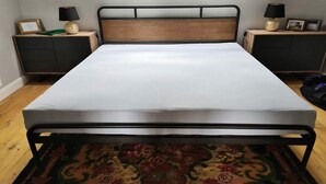 Cama de solteiro com estrutura metálica e cabeceira de madeira, acompanhada por duas mesinhas de cabeceira com abajures, em um quarto com piso de madeira.