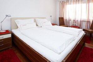 Cama bem arrumada com lençóis e almofadas brancas com a inscrição 'Zeitlos' em um quarto de hotel, com cabeceira de madeira, tapete vermelho e janela com cortinas listradas.