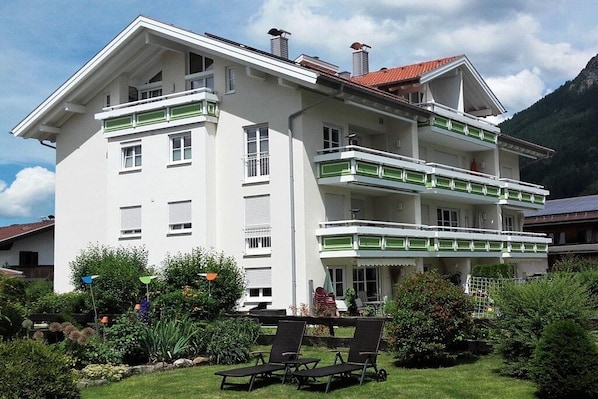 Um edifício de vários andares com varandas, um jardim com área de estar ao ar livre e montanhas ao fundo.