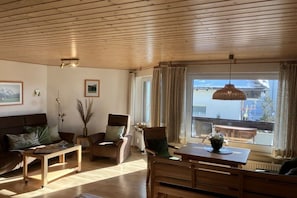 A living room with a wooden ceiling, a sofa, a chair, a table, and a view of a building through a window. - Alpenflair Holiday Apartment 102 (Oberstdorf)