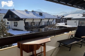 A balcony with a wooden table and a chair, overlooking a residential area with houses and mountains in the background. - Alpenflair Holiday Apartment 102 (Oberstdorf)