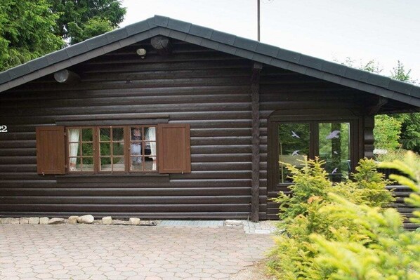 Eine Blockhütte mit Satteldach, einem Fenster mit Holzläden und einem gepflasterten Eingang.