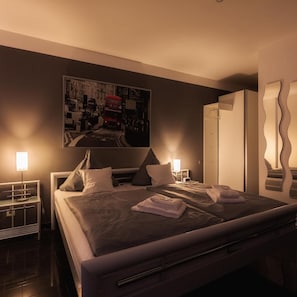 A modern hotel room with a large bed, bedside tables, a wardrobe, and a cityscape painting on the wall. - Hotel Ruhen (Rühen)