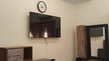 A modern living room with a flat-screen TV mounted on the wall, a wooden cabinet with drawers, a small refrigerator, and a large mirror.