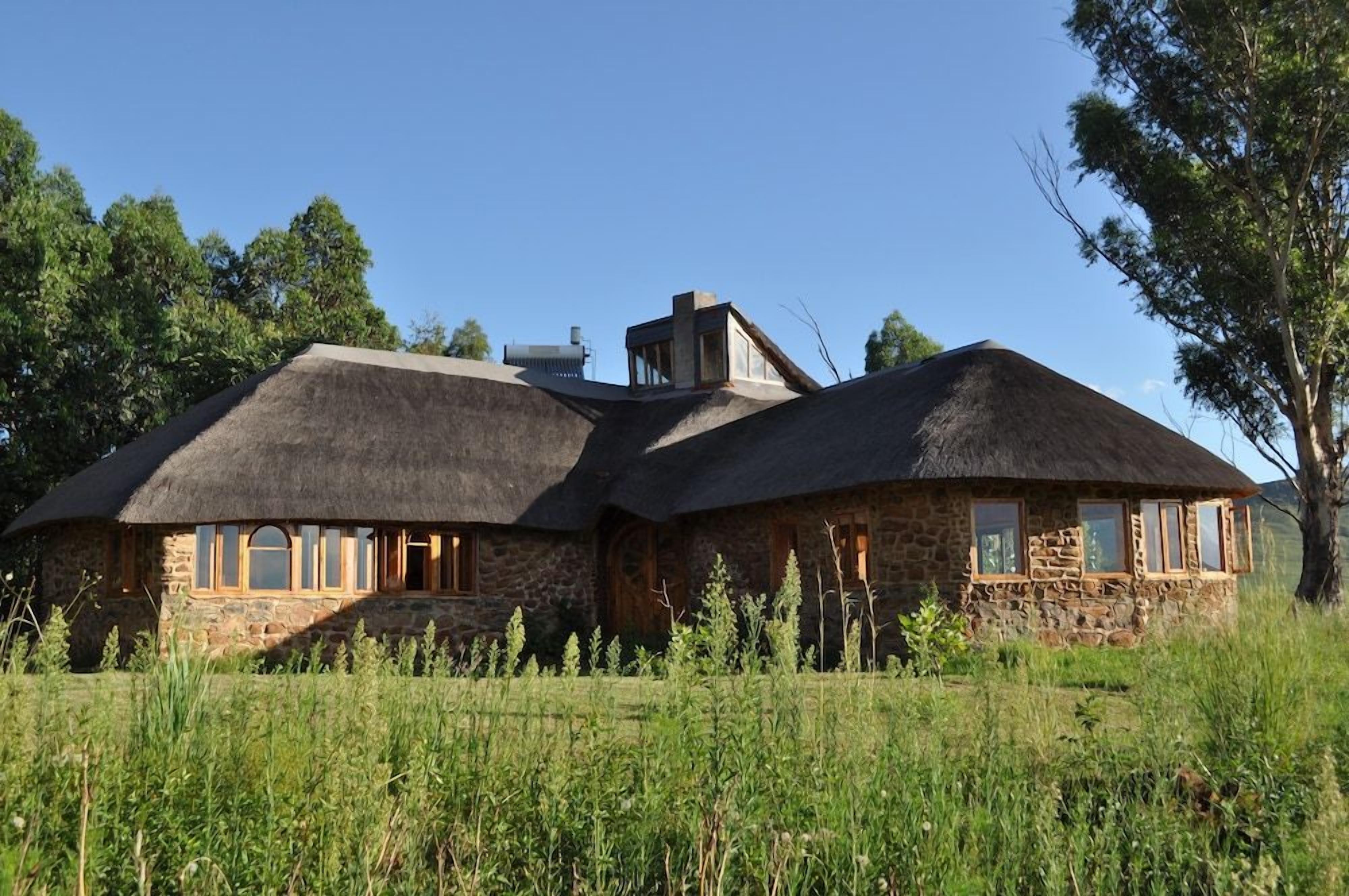 A stone house with thatched roofs and a chimney, surrounded by a grassy field and trees.