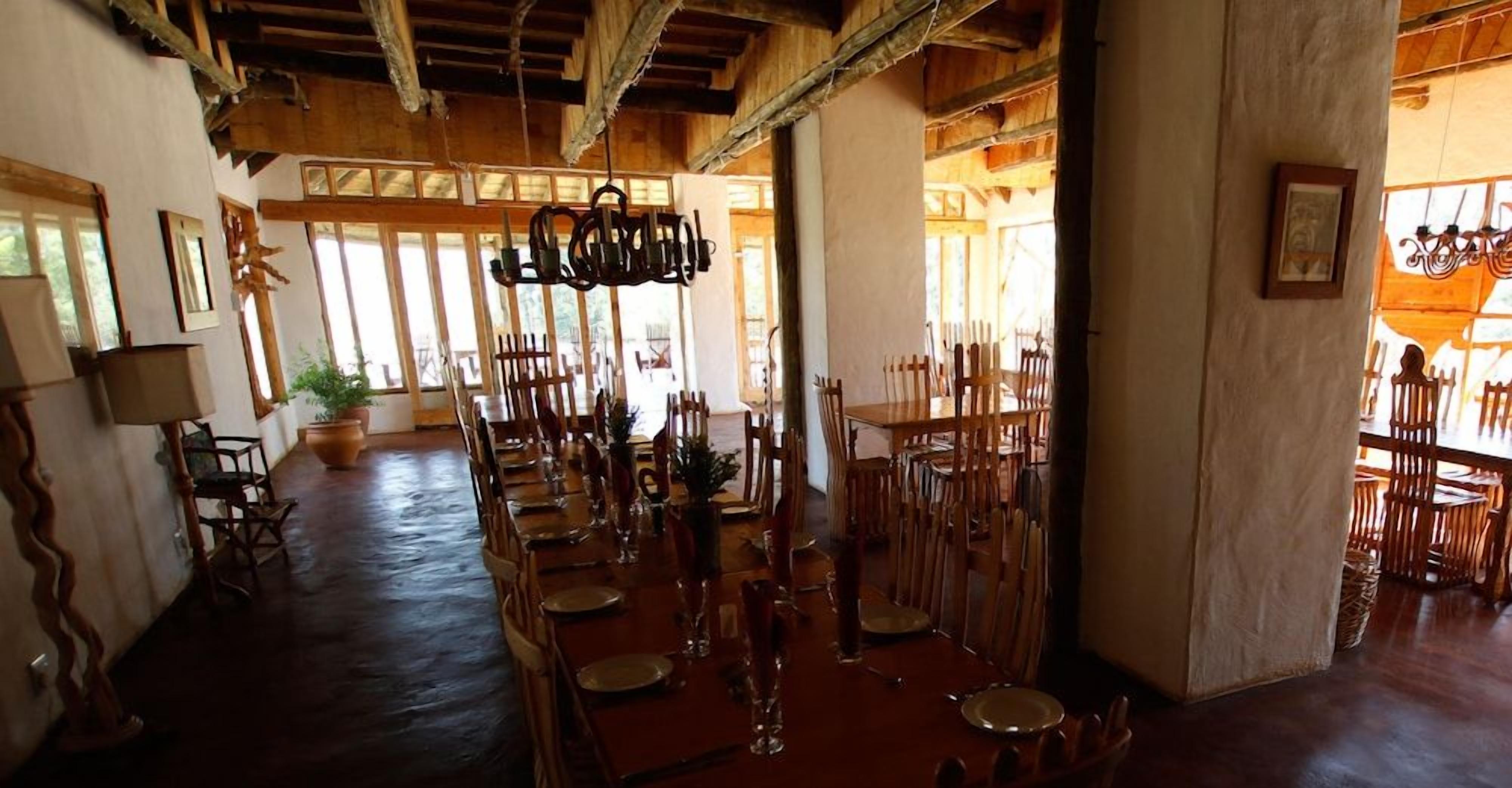 A dining area with wooden tables and chairs, a chandelier, and large windows allowing natural light.