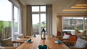 A dining area with a wooden table set for four, a lantern, and a view of a garden through large windows. - Mastiff Select Riveria Resort, Igatpuri Road, Nashik (Nashik)