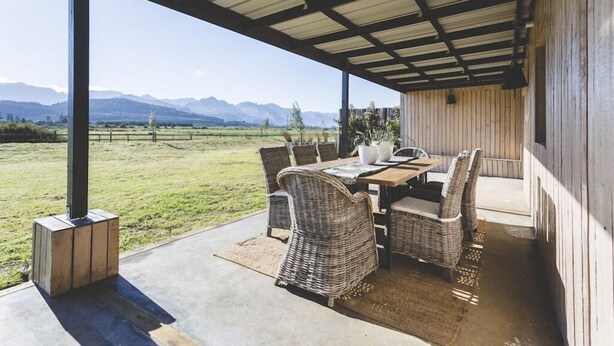 Un patio al aire libre con una mesa y sillas, un área cubierta y vistas a un campo de césped y montañas.