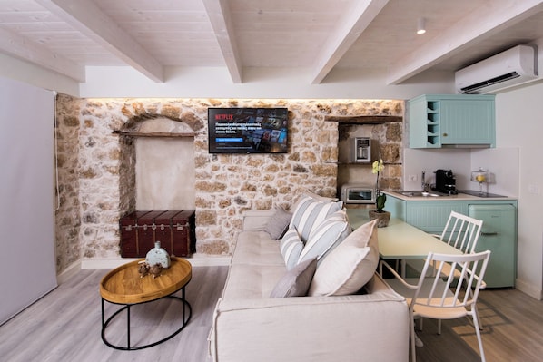 Una zona d'estar acollidora amb una paret de pedra, televisió de pantalla plana, sofà, taula de cafè i una cuina amb un rentaplat i armaris.