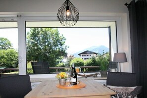 A room with a large window, a wooden table with a bottle and flowers, and a chair. - Alpenflair Holiday Apartment 107 (Oberstdorf)