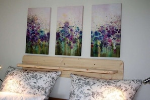 A bed with a wooden headboard and floral-patterned pillows, flanked by three abstract paintings on the wall. - Alpenflair Holiday Apartment 107 (Oberstdorf)