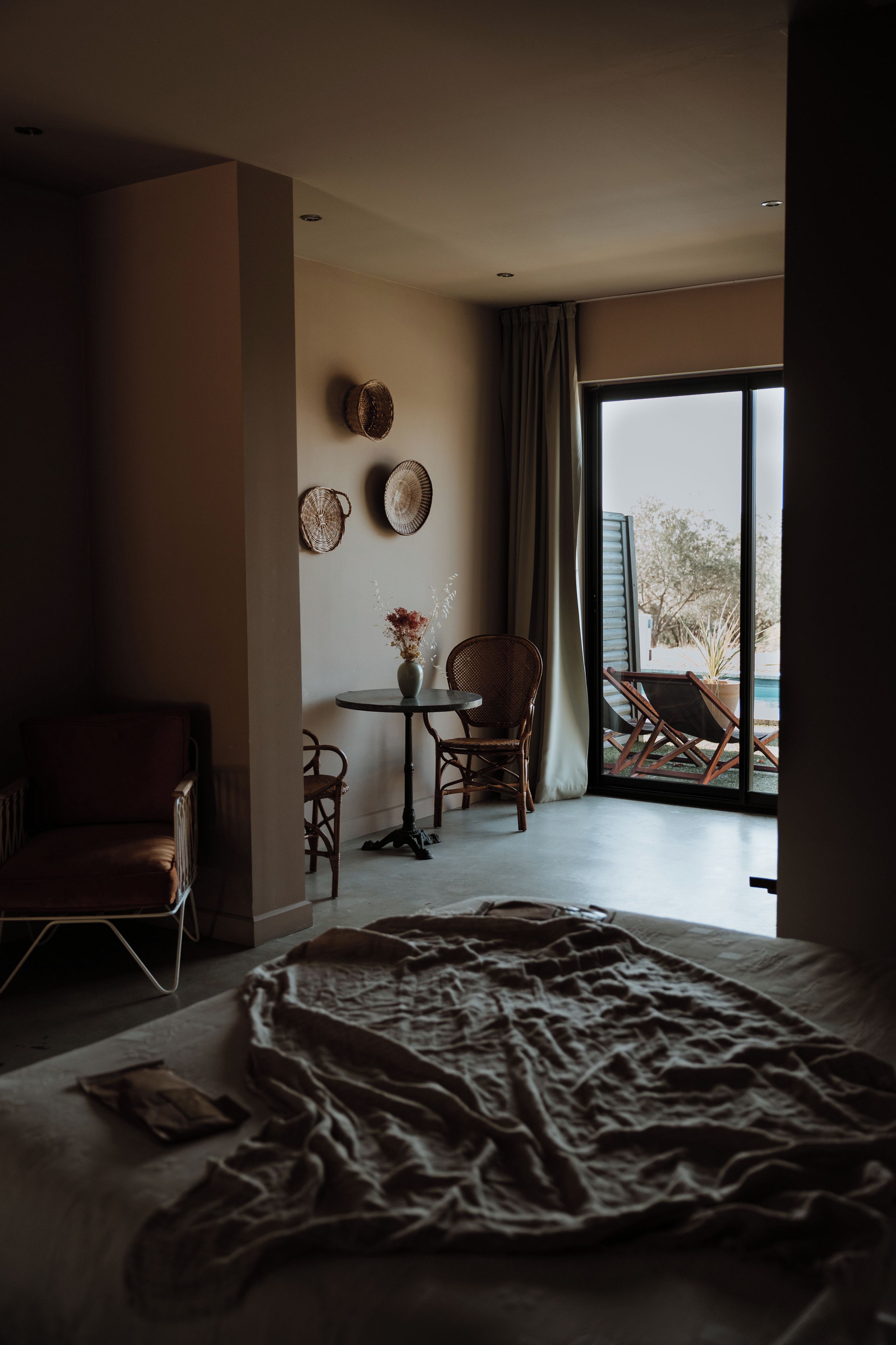 A bedroom with a large bed, a small table with a vase, two chairs, and a view of the outdoors through a large window.