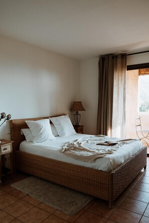 A room with a wicker bed, a nightstand, a lamp, a chair, and a view through a window with curtains. - Hotel Auberge La Feniere (Cadenet)
