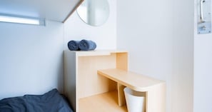 Une pièce minimaliste avec un lit, une étagère en bois et une poubelle blanche.