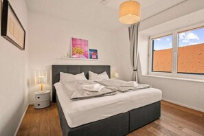 Une chambre à coucher avec un lit, des lampes de chevet, une fenêtre avec des rideaux et des œuvres d’art accrochées au mur.