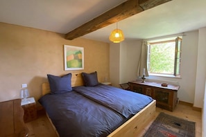 A bedroom with a wooden bed, a bedside table, a nightstand, a window with curtains, and a hanging light fixture.