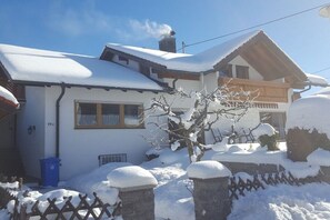 Uma casa coberta de neve com varanda de madeira, chaminé visível e lixeira azul.