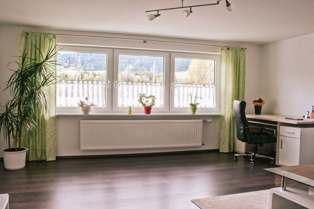 A room with a large window, a radiator, a desk, a chair, and a potted plant.
