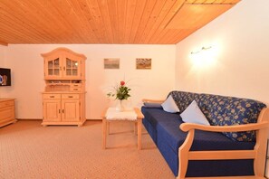 A cozy living room with a wooden ceiling, a blue sofa with floral cushions, a wooden cabinet, and a small table with a vase of flowers. - Hefle in the Gottstein House (Bad Hindelang)