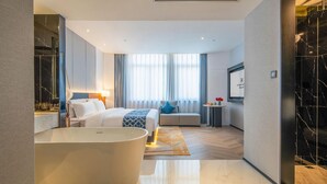 Quarto de hotel moderno com uma banheira grande, cama com colcha azul estampada e um espaço de estar com um almofada azul.