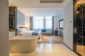 Habitació d'hotel moderna amb una banyera gran, un llit amb un estriat de color blau i una zona de seients amb un coixí blau.