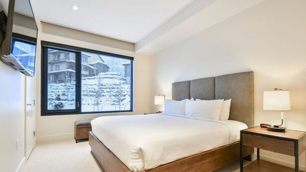 Un dormitorio moderno con una cama grande, mesitas de noche, un ventanal con vistas a un paisaje nevado y un televisor montado.