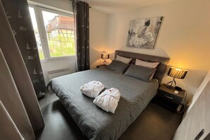 A hotel room with a bed, two bedside lamps, a painting on the wall, and a window with curtains. - Holiday apartment with 1 bedroom (COLMAR)