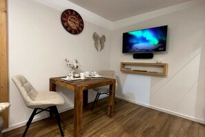 Ein Zimmer mit einem Holztisch, zwei Stühlen, einer Wanduhr, einem Fernseher und einem Regal.