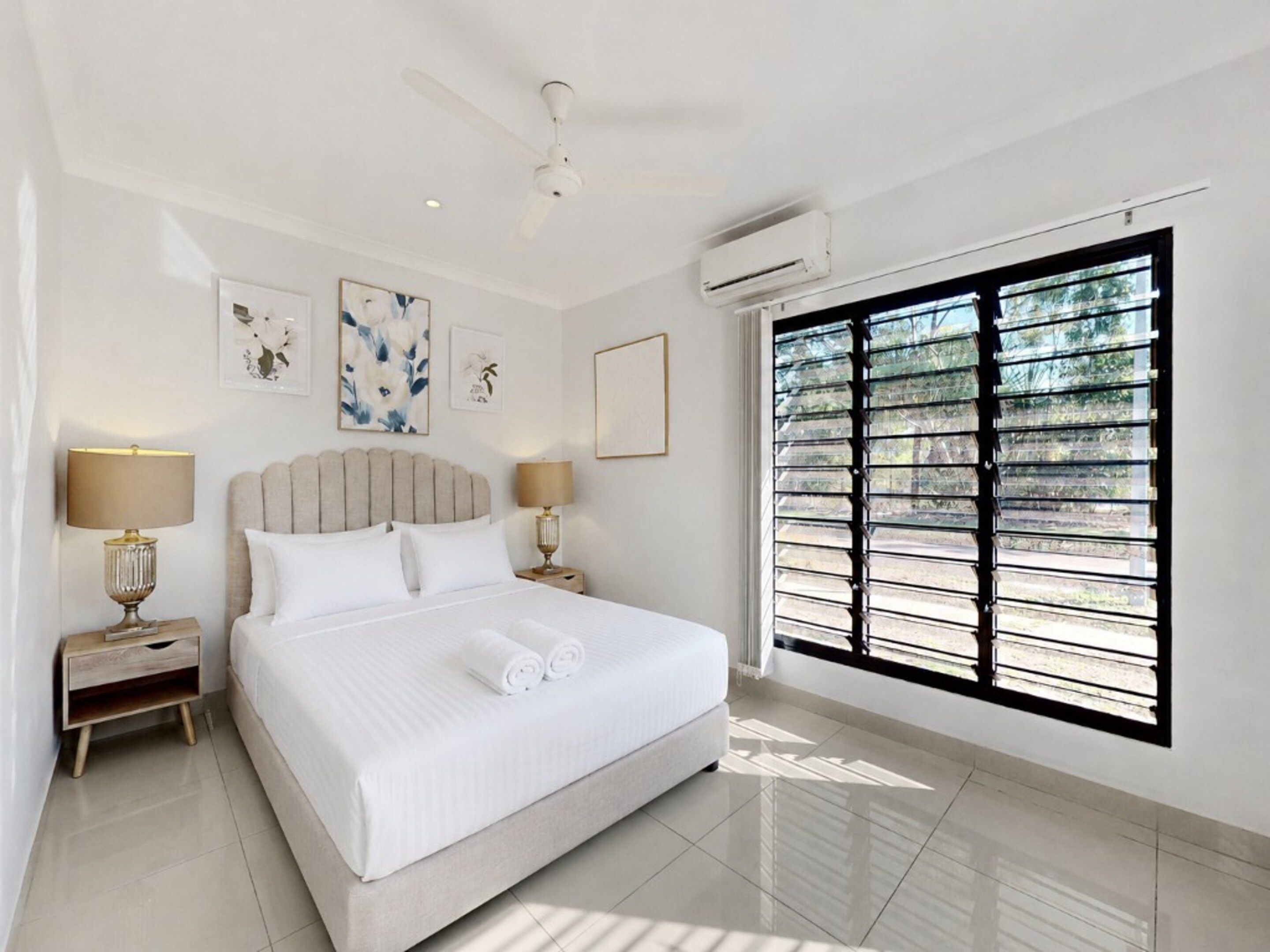 A hotel room with a large bed, two bedside tables, a ceiling fan, an air conditioning unit, and a window with blinds.