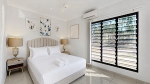Une chambre d’hôtel avec un grand lit, deux tables de chevet, un ventilateur de plafond, un appareil de climatisation et une fenêtre avec des stores.