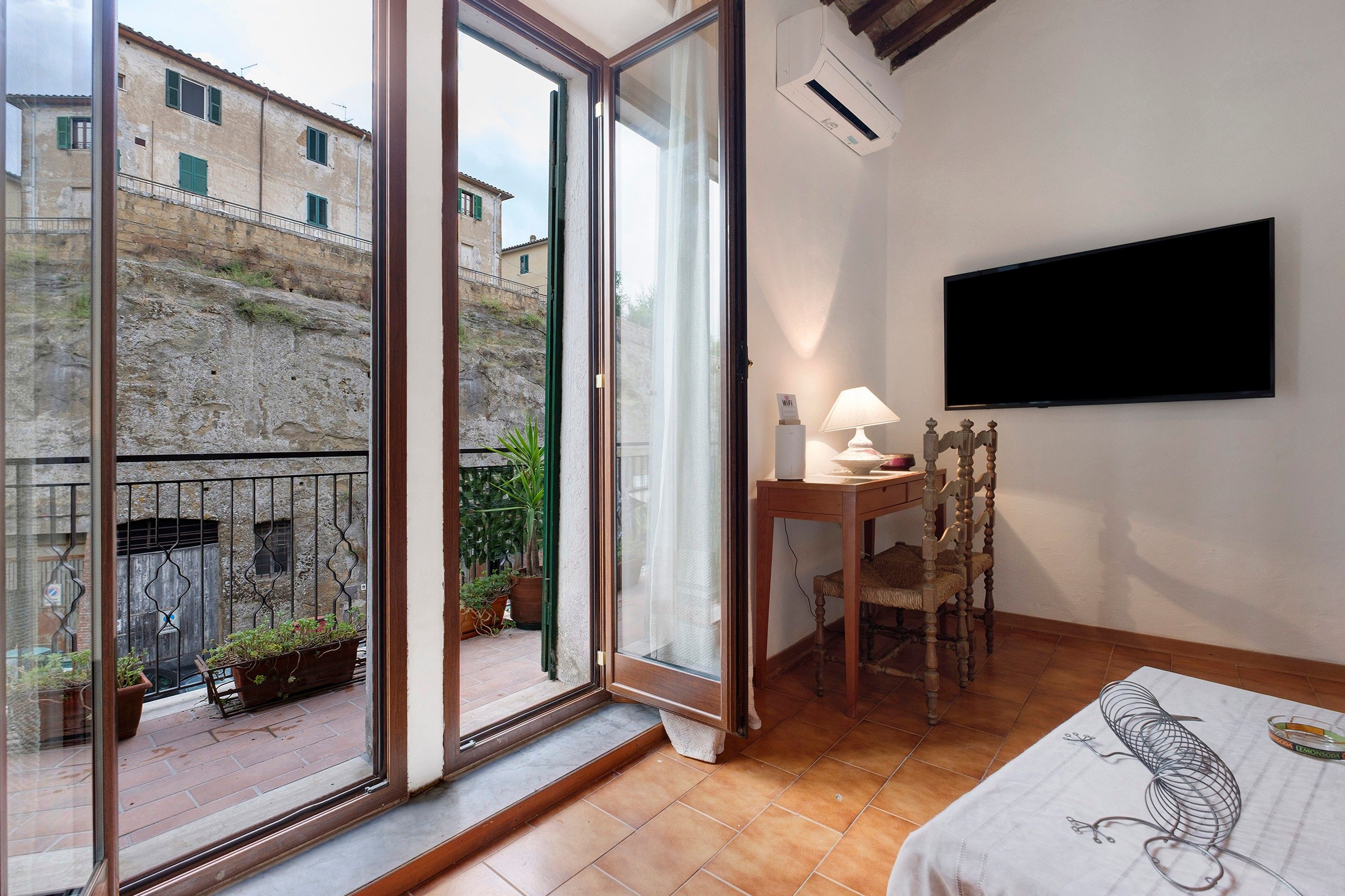 Ein Zimmer mit einem Bett, einem Tisch mit Lampe, einem Fernseher und einem Balkon mit Pflanzen.