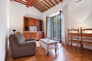 A cozy living room with a sofa, a dining table, and a wooden chair. There's a kitchen with wooden cabinets and a window with a view.