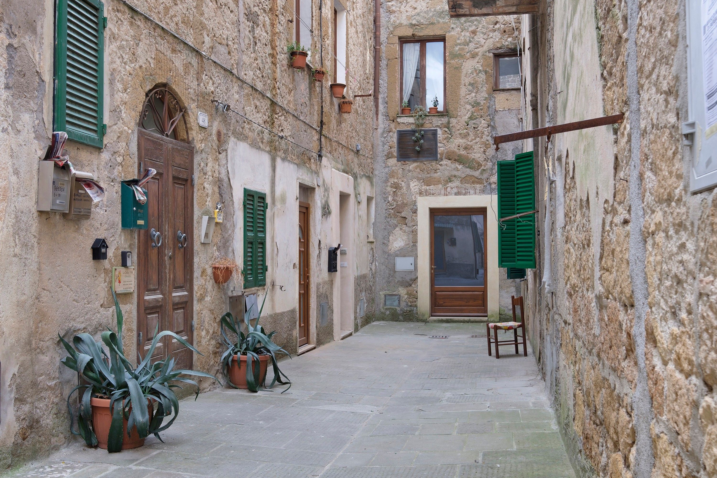 Un viuzza stretta con pareti in pietra, porte di legno e persiane verdi. Si vedono piante in vaso e una sedia.
