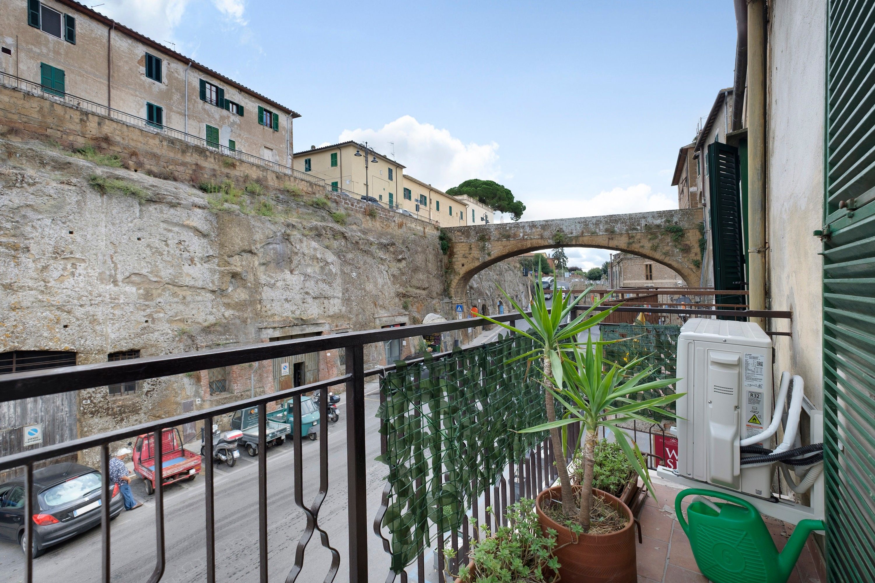 A balcony with a view of a stone wall, a street with cars, and a building with a green awning.