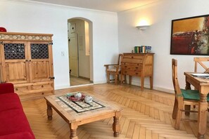 Une pièce avec un parquet en chevrons, des meubles en bois, un canapé rouge, un buffet en bois doté de portes en verre, un bureau en bois avec une chaise, et un tableau accroché au mur.