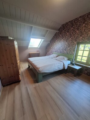 A bedroom with a sloped ceiling, a skylight, a bed with white linens, a wooden dresser, and a floral-patterned wall. - Vrbo Property (Aarle-Rixtel)