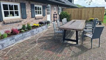 A brick house with a wooden deck, a dining table with chairs, and a garden with various plants.
