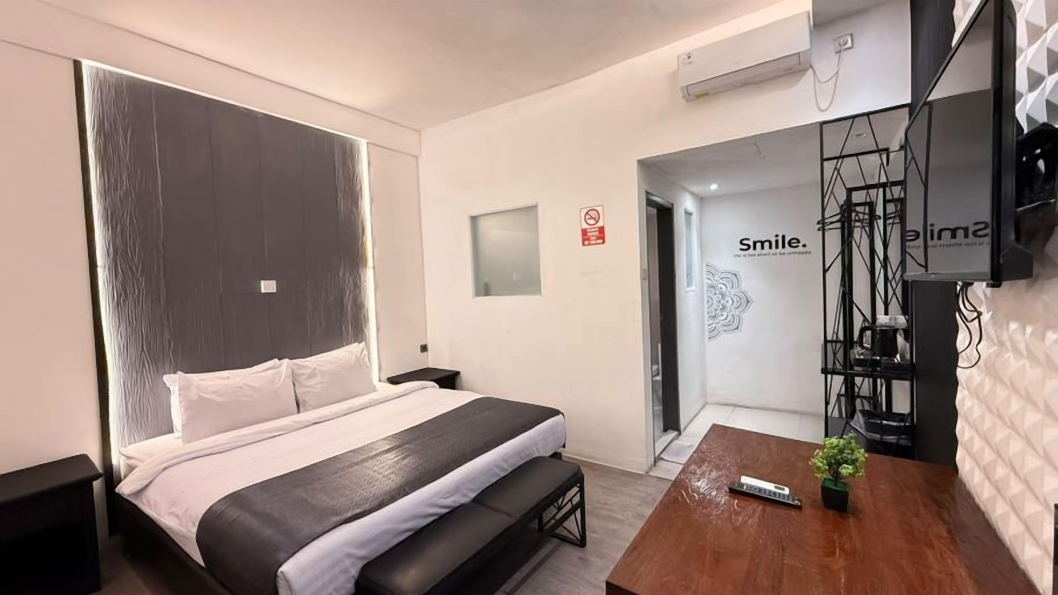 A modern hotel room with a large bed, a small desk, a TV, and a wall with a 'Smile' sign.