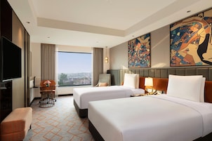 Habitación de hotel con dos camas, televisión, escritorio con silla, ventana con cortinas y obras de arte en las paredes.