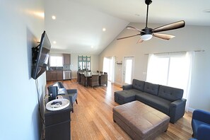 A spacious living area with a large gray sectional sofa, a flat-screen TV mounted on the wall, a ceiling fan, and a dining area with a glass table and wicker chairs. - Amazing  Beach House with steps to the beach, Ocean views! (North Topsail Beach)
